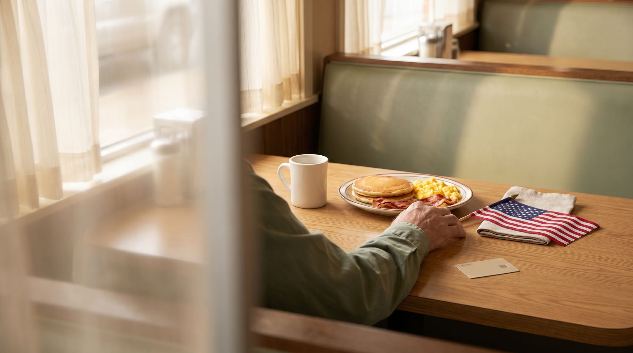 A veteran's hand in a sage green sleeve rests on a diner table beside a plate of pancakes, eggs and bacon, a white coffee mug, a small American flag on a linen napkin and a blank ID card.