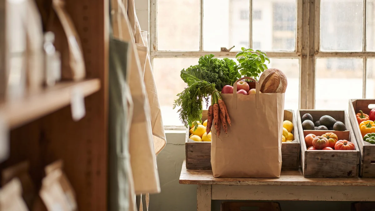 A paper bag of fresh vegetables, fruit and bread on a wooden table beside crates of produce in a grocery store.